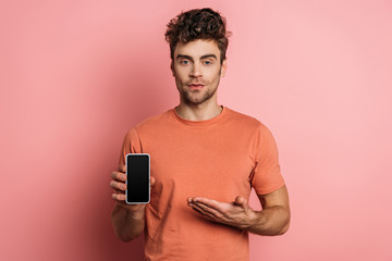 serious young man pointing with hand at smartphone with blank screen on pink background