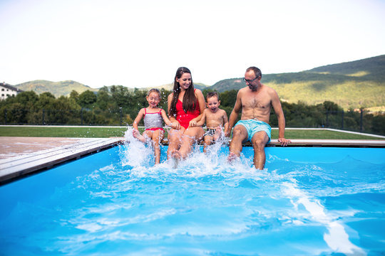 Young Family With Two Small Children Sitting By Swimming Pool Outdoors.