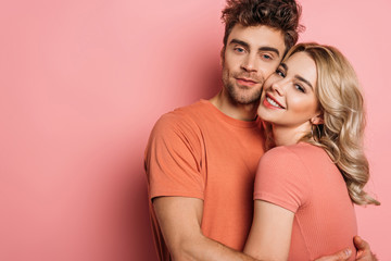 happy young couple embracing while looking at camera on pink background