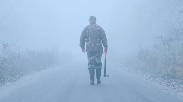 Steadicam shot of Strange man with an axe walks down the road in the fog. Back view