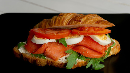Salmon sandwich with tomato, egg and soft cheese in croissant on black plate on a white wooden table