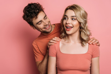 cheerful man touching shoulders of flirty girl on pink background