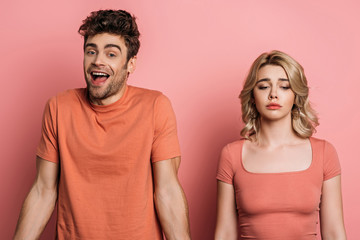 cheerful man looking at camera while standing near offended girlfriend on pink background
