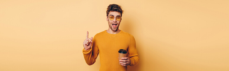 panoramic shot of excited man showing idea sign while holding disposable cup on yellow background