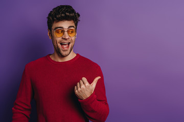 cheerful young man pointing with thumb and looking away on purple background