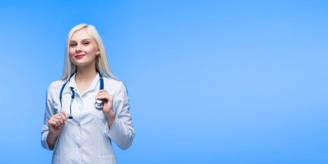 Medical concept of beautiful female doctor in white coat with phonendoscope. Medical student general practitioner. Woman hospital worker looking at camera and smiling, studio, Blue background