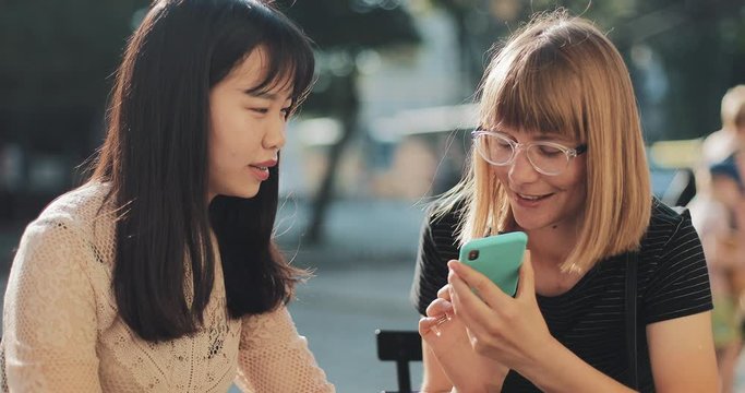Two girls sitting outside and using smartphone voice recognition. Cheerful girl speaking to foreigner friend through smartphone voice translator application. Concept of communication and tech.
