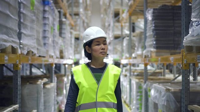 Front view of young asian woman walking on warehouse interior during working day. Female worker inspects shelves with industrial products, doing work at factory storehouse. Employee wearing helmet and