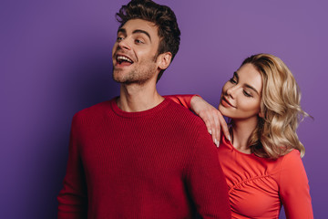 happy girl leaning on shoulder of smiling boyfriend looking away on purple background