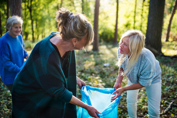 Senior women friends picking up litter outdoors in forest, a plogging concept.