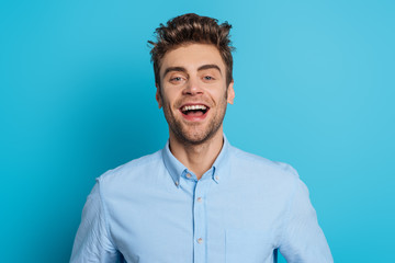 cheerful young man laughing while looking at camera on blue background