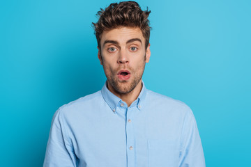 Fototapeta premium shocked young man looking at camera while standing with open mouth on blue background