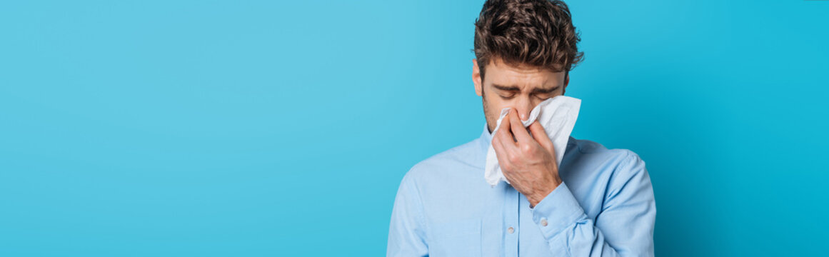 panoramic shot of diseased man sneezing in paper napkin on blue background