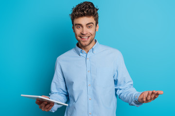 cheerful young man standing with open arm while holding digital tablet and smiling at camera on blue background