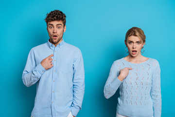 shocked man and woman pointing with fingers at themselves while looking at camera on blue background