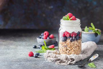 Granola Parfait with Yogurt, oat granola, fresh berries, honey and mint leaves in tall glass jar, copy space