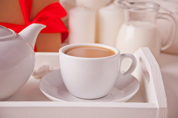 Cup of coffee, teapot on the white tray with gift box and candles nearby.