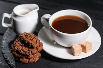 Coffee with milk in an white cup, served with biscuits and sugar on a dark background.