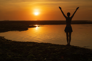 Excited young woman raising arms at the beach in front of the ocean. View from back. Sunset golden hour at the beach. Bali, Indonesia.