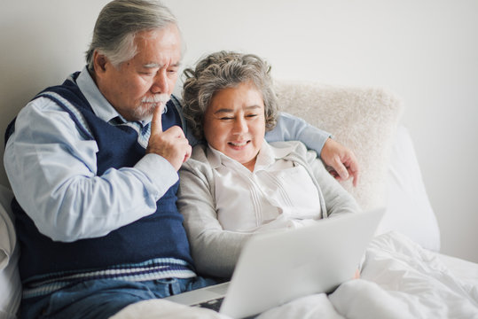 Senior Asian Couple Siting On The Bed And Looking At Computer Laptop For Health Check Report, Retired Man And Woman Using Notebook To Video Call Which Smiling And Felling Happy In Bed Room At Home.