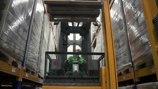 Caucasian man worker loads shelves while sitting in loading machine at industrial warehouse. Male operator is doing work in storehouse of plant, managing modern equipment. Employee wearing uniform