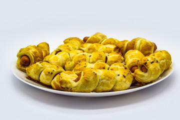 Baked ausages in the dough on a white plate against white background. Sausage Puff Pastry Buns.sausages wrapped in puff pastry and baked on the plate. A tasty mini buns with sausages.