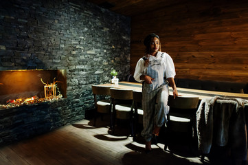 African american woman in overalls and beret in room with wooden table and fireplace.