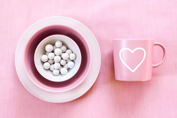 Pink mug and plate with white sugar dragee on gentle pink cloth background. Top view, flat lay