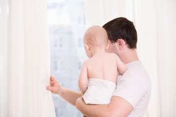 Young man stands looking through curtains with toddler son