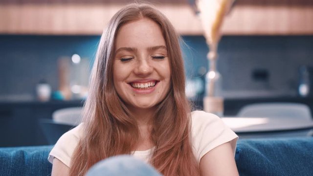 Portrait of young attractive red hair woman in white t-shirt sitting in blue coach looking to camera and smiling in modern apartment feeling happy great evening good emitions home comfortable