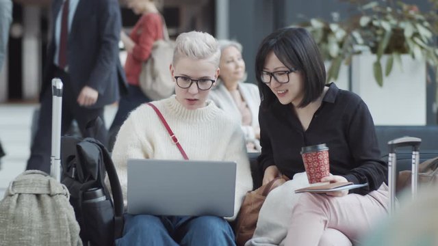 Medium pan shot of two young Caucasian and Asian girlfriends with luggage sitting in departure lounge at airport, looking at laptop screen and chatting, and multinational passengers waiting for flight