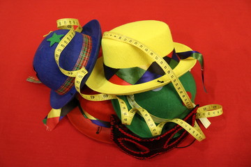 A yellow serpentine-wrapped measuring tape is laid on top of multicolored hats and a masquerade mask on a red background. Side view.  
