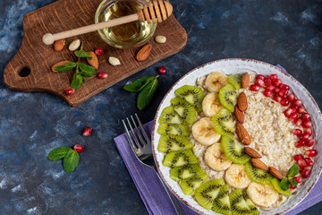 Breakfast consisting of oatmeal, nuts and fruits. Kiwi orange pomegranate almonds bananas decorate a plate. Healthy eating, on a dark background. Copy space.