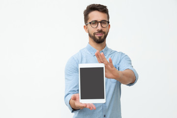 Confident man showing digital tablet with blank screen. Front view of handsome bearded young man in eyeglasses holding tablet pc with blank screen and looking at camera. Advertisement concept