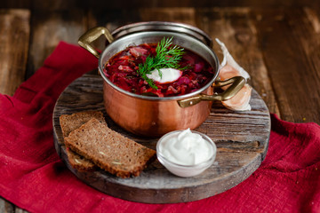 Freshly cooked homemade russian famous soup borsch. Served in cooper pan with garlic, sour cream and dill. Nourishing tasty meal. Rustic style, close up, wooden background, red tissue as decor. 