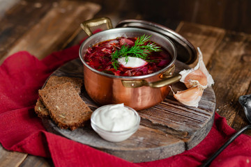 Freshly cooked homemade russian famous soup borsch. Served in cooper pan with garlic, sour cream and dill. Nourishing tasty meal. Rustic style, close up, wooden background, red tissue as decor. 