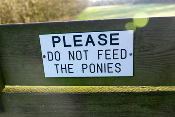 Please do not feed the ponies sign attached to a wooden board on a style with  field in background