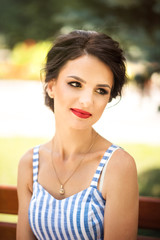 Fototapeta premium Vertical close up portrait of young beautiful brunette woman in blue and white striped dress with makeup and red lipstick looking away in park in summer. Summer is allergy time