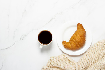 Cup with coffee, croissant on a plate and a knitted scarf on a marble background. French breakfast concept. Flat lay, top view