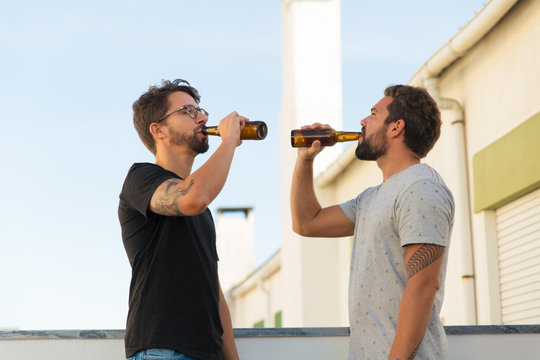 Side view of two handsome young man drinking beer. Friends with beer bottles hanging out on balcony. Leisure, friendship concept