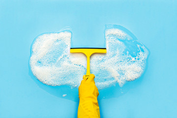 Hand in a yellow rubber glove holds a scraper for cleaning and wipes soap suds on a blue...