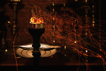 Shisha hookah bowl with red hot coals and craft tobacco. Modern hookah with coconut charcoal for relax and smoke. Beautiful sparks in long exposure on cozy dark lounge background.