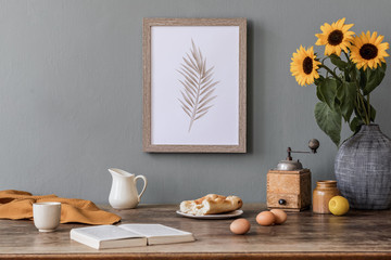 Stylish and sunny interior of kitchen space with wooden table, brown mock up photo frame, breakfast and sunflowers. Scandinavian home decor with kitchen accessories. Template. Gray color concept