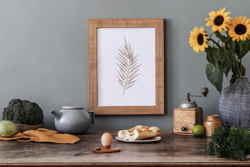 Stylish and elegant home interior of kitchen space with wooden mock up poster frame, sunflowers in vase, fresh vegetables and kitchen accessories. Nice home decor. Template. Gray color concept.