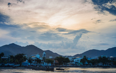 Sunset over Ubatuba beach, with calm waters in the foreground and the city and lush mountains in the background under a clear sky with some clouds