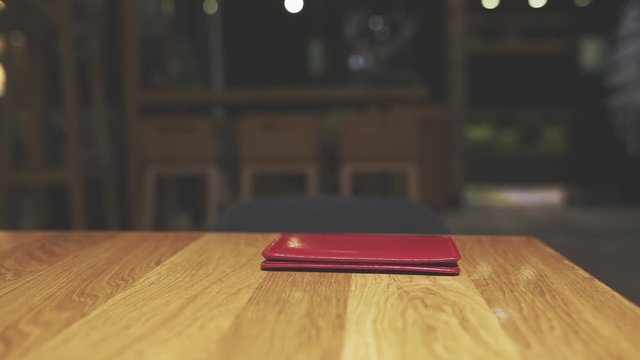 Red leather bill folder lying on brown wooden table in cafe or restaurant. Unidentified waiter passing by on blurry unfocused interior background.