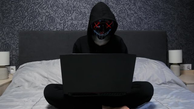Female hacker dressed in black mask and hoodie working on laptop in the bedroom, woman hacking and hacker attack