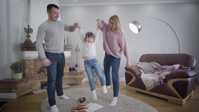 Young Caucasian husband and wife holding daughter's hands and raising child up. Playful positive family enjoying time together indoors. Unity, lifestyle, happiness.