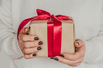 A woman wearing a knitted white sweater holds a gift box with a red ribbon.