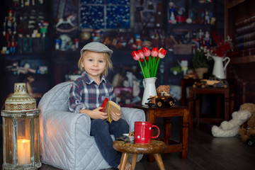 Sweet toddler boy, wearing glasses, reading a book and drinking tea with cookies